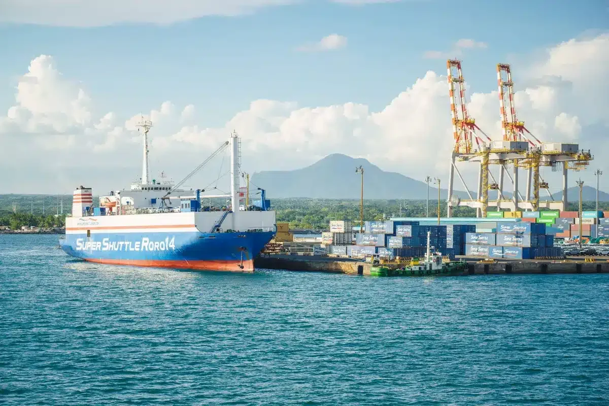 A cargo ship docked at an industrial port with shipping containers and cranes under a clear sky.