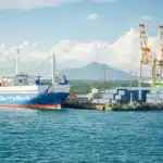A cargo ship docked at an industrial port with shipping containers and cranes under a clear sky.