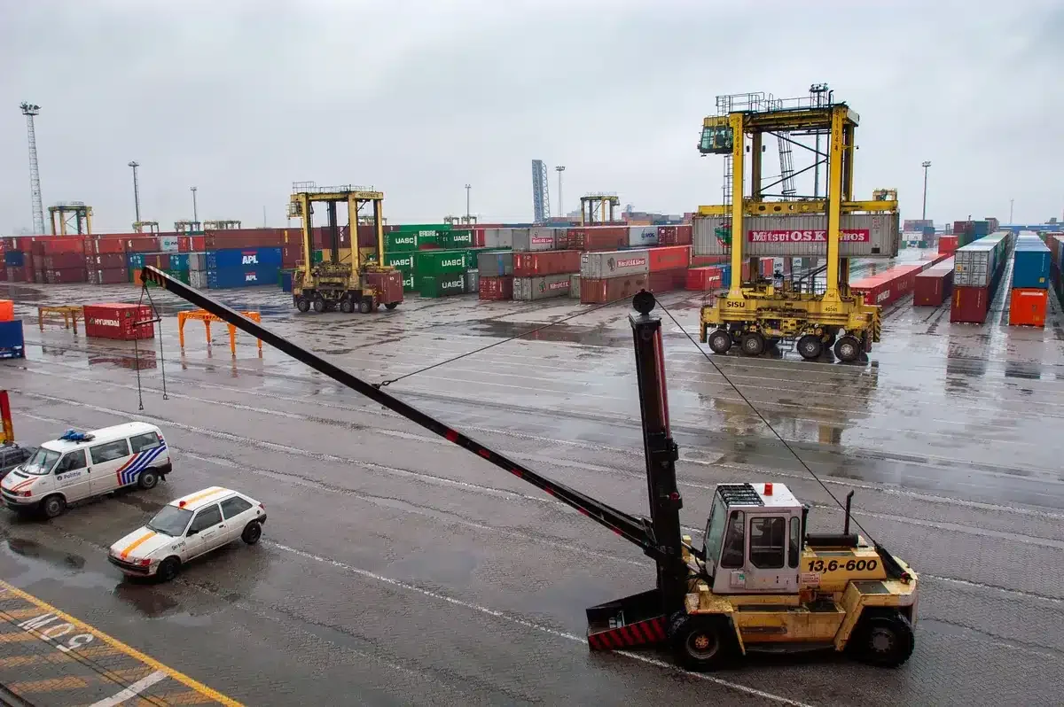Container terminal with straddle carriers and stacked shipping containers under a cloudy sky.