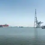 A large container ship sails through an industrial harbor under a clear sky, showcasing maritime log