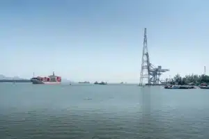 A large container ship sails through an industrial harbor under a clear sky, showcasing maritime log