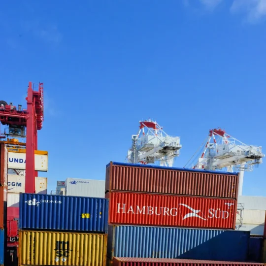 Colorful cargo containers stacked at a busy shipping port under a clear blue sky.